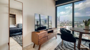 Een moderne hotelkamer met een grote spiegel, een flatscreen televisie, een houten dressoir, een bureau en uitzicht op een stadsbeeld door grote ramen.