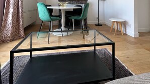A modern living room with a glass coffee table, a dining table with chairs, a large mirror, and a floor lamp.