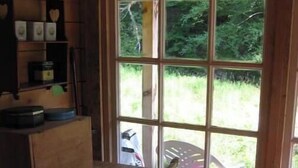 Interior rústico de una cabaña con una mesa y silla de madera, una ventana que da a la vegetación y una alfombra estampada en el suelo.