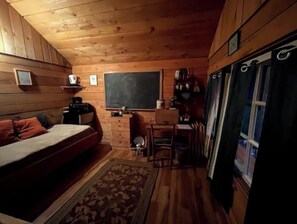 A cozy cabin room with a bed, a desk, a chair, a small rug, and a window with curtains. - Farm Stay in Catskills FarmHand Cabin (East Meredith)