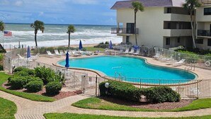 A pool area with a view of the ocean, surrounded by a fence, and a building with balconies. - Bent Palm 202 | Oceanfront Condo with Balcony Views, Pool + Beach Access (Ormond Beach)