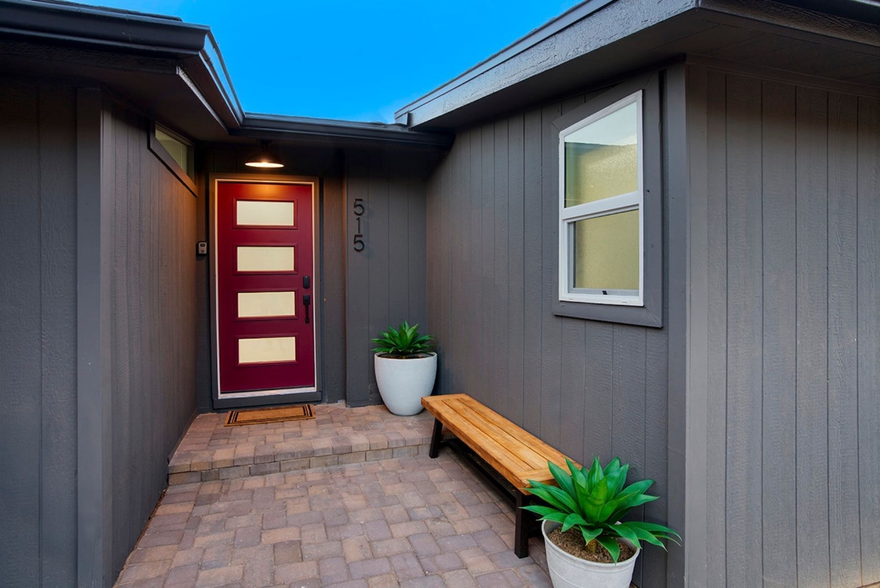 Une maison avec une porte rouge, un banc et deux plantes en pot.