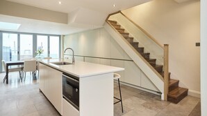 Uma cozinha moderna com uma ilha central, uma área de jantar com mesa e cadeiras, e uma escadaria com degraus de madeira e guarda-corpo de vidro.