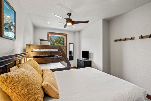 A bedroom with a bunk bed, a ceiling fan, a television, and a window with a view of trees. - Stylish, Inviting Townhome~Near Skiing~Hot Tub+Pool (Steamboat Springs)