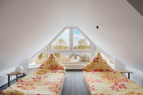 A bedroom with a sloped ceiling, a large window, a bed with floral bedding, and a small table with a lamp.