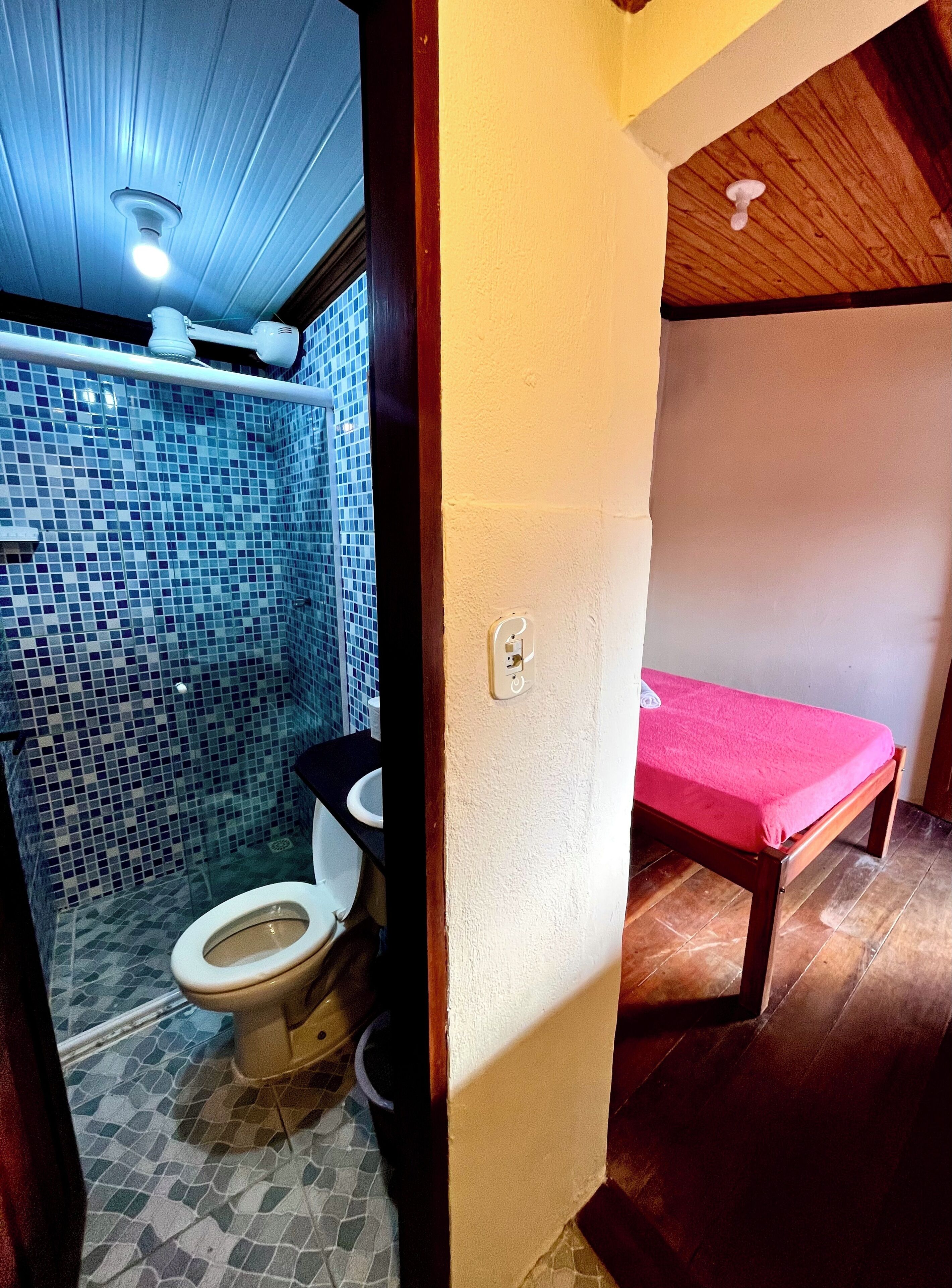 A bathroom with a toilet, a shower area with blue mosaic tiles, and a wooden bench with a pink towel.