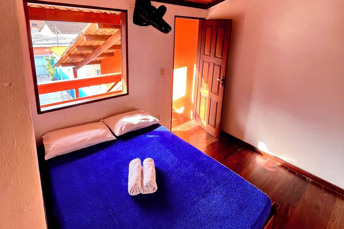 Un lit simple avec un matelas bleu, des oreillers blancs et une paire de serviettes enroulées. Un ventilateur de plafond est visible au-dessus du lit, et une fenêtre offre une vue sur l’extérieur.