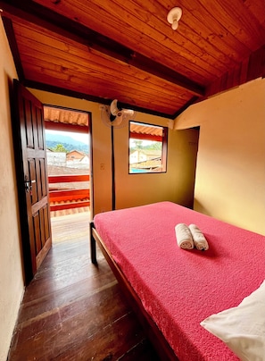 A room with a wooden ceiling, a bed with a red bedspread, two rolled towels, and a view of a mountainous landscape through a window. - Pouso Paraty (Paraty)