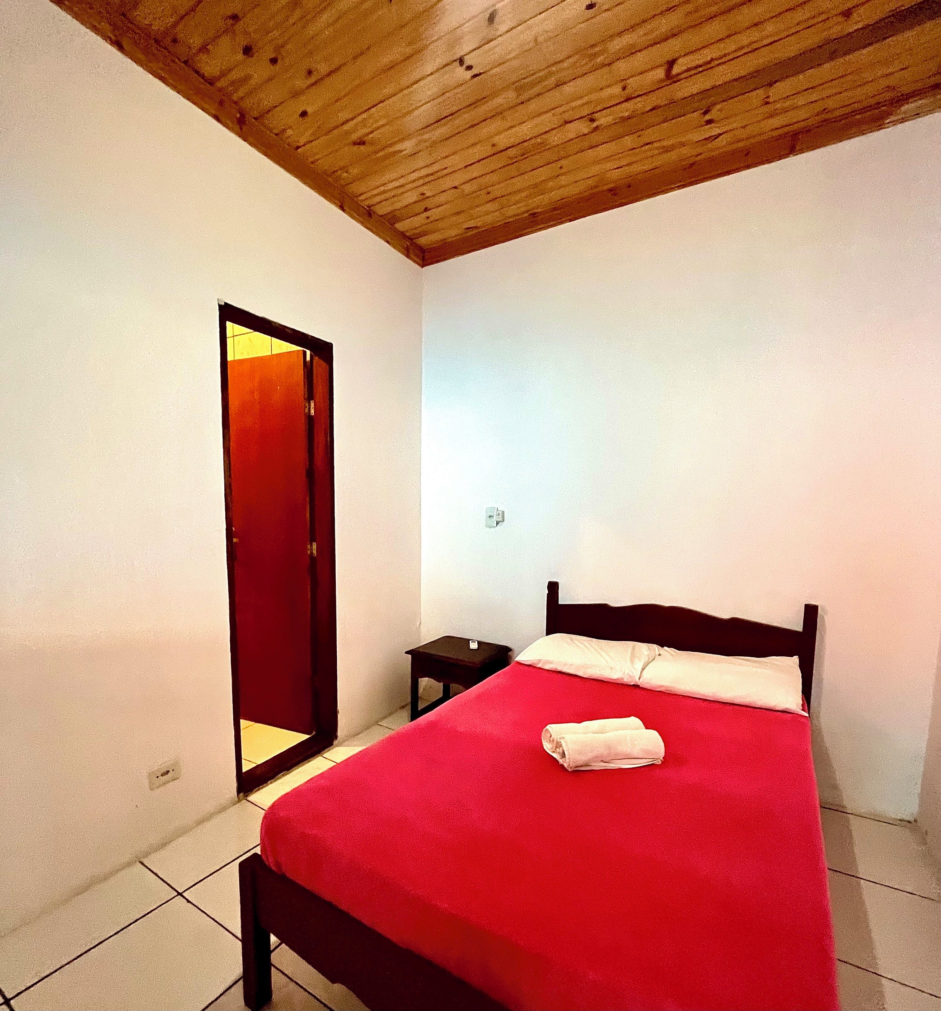 A bedroom with a wooden ceiling, a bed with a red bedspread, a small bedside table, and a door leading to another room.