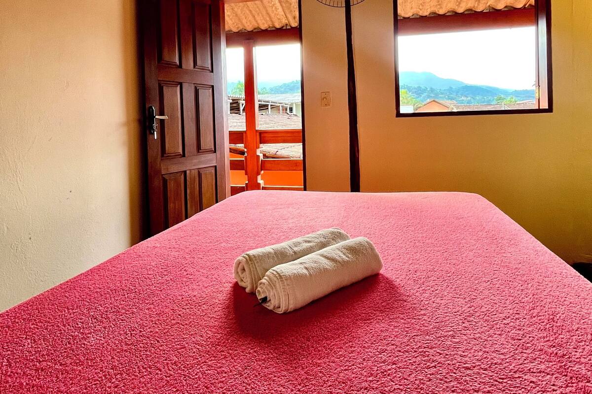Une chambre avec un lit recouvert d’une moquette rouge, un plafond en bois, un ventilateur et une fenêtre donnant sur les montagnes.