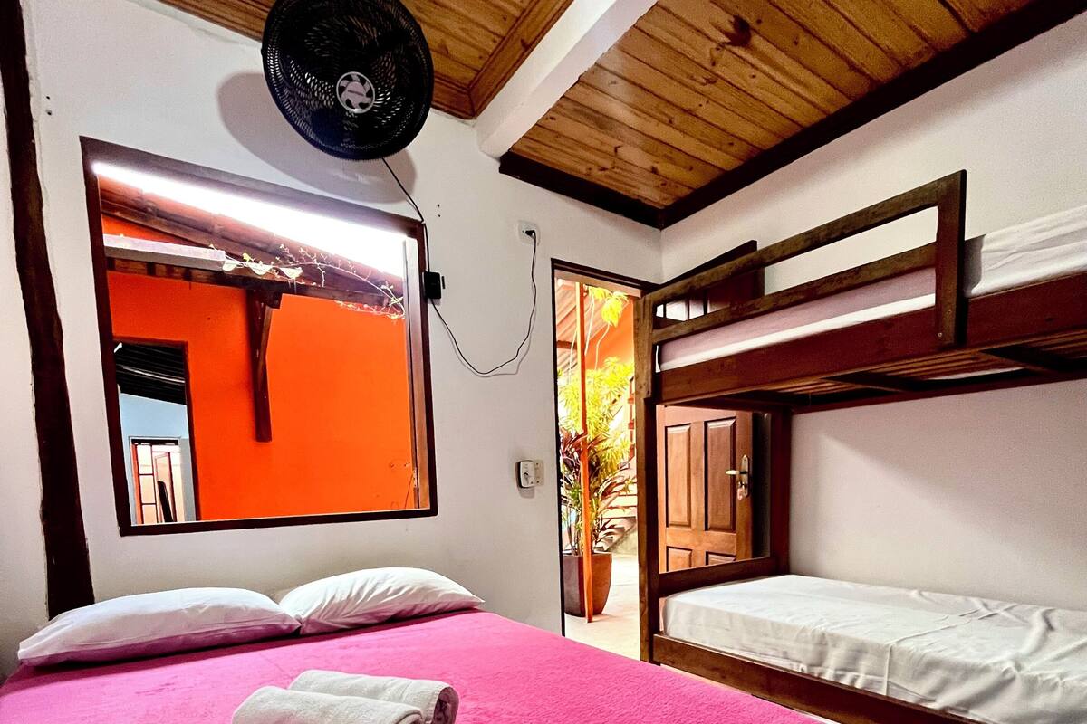 Une chambre avec un lit superposé, un lit simple avec une couvre-lit rose, une fenêtre donnant sur un paysage orangé et un ventilateur de plafond.