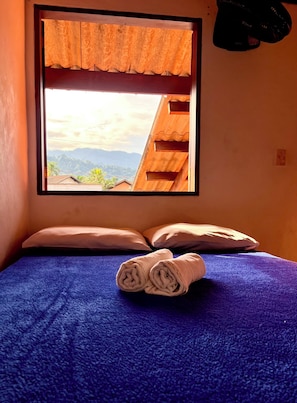 Een kamer met een bed, kussens, een opgerolde handdoek, een raam met uitzicht op heuvels en een houten plafond.