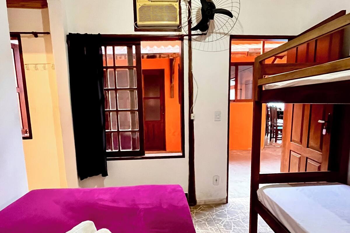 Une chambre avec un lit superposé, un lit recouvert d’une couverture violette, un ventilateur et un appareil de climatisation.