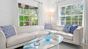 A modern living room with a glass coffee table, two sofas with patterned cushions, and large windows with a view of greenery.