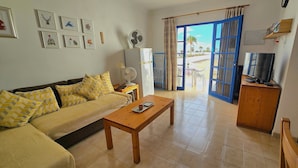 A living room with a sofa, coffee table, and a television on a wooden cabinet. There are framed pictures on the wall and a fan on a table. - Atalaya Complex, central Puerto del Carmen, sunny terrace with sunloungers (Tías)