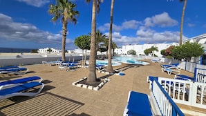Ein Poolbereich mit Liegestühlen und Palmen, ein Schwimmbecken sowie Meer- und Stadtblick im Hintergrund.