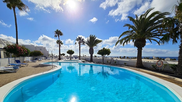 Ein Swimmingpool mit klarem, blauem Wasser, umgeben von Palmen und Liegestühlen, mit Blick auf das Meer im Hintergrund.