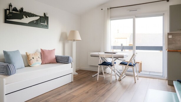 Un salon moderne avec un canapé blanc, une table à manger et des chaises. Un tableau représentant un paysage urbain orne le mur, et une fenêtre offre une vue sur la ville.