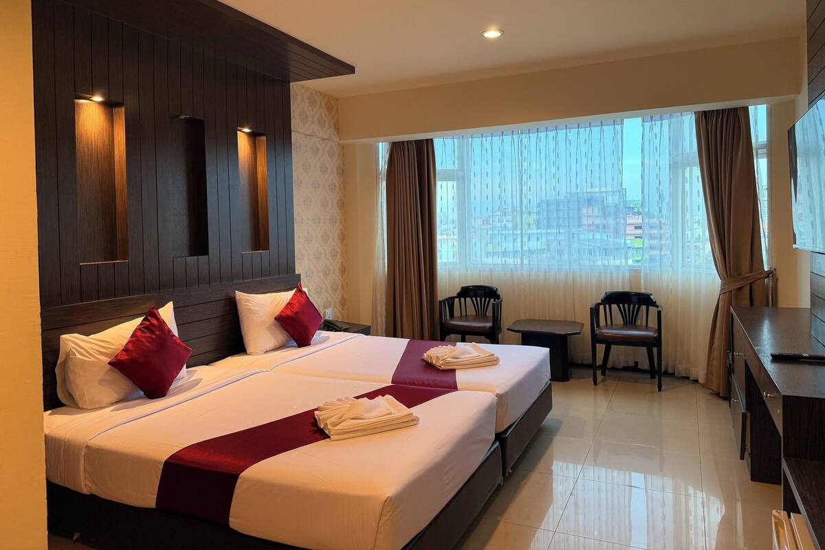 A hotel room with two beds, a desk, chairs, and a large window with curtains.