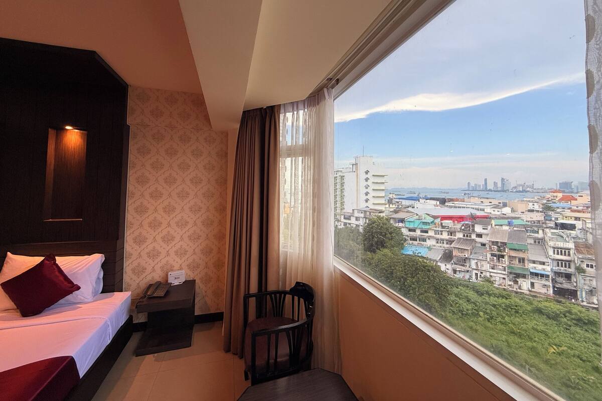 A hotel room with a bed, a desk, a chair, and a large window offering a city view.