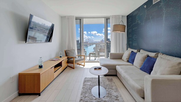 A modern living room with a sofa, a coffee table, a TV mounted on the wall, and a view of a cityscape through large windows.