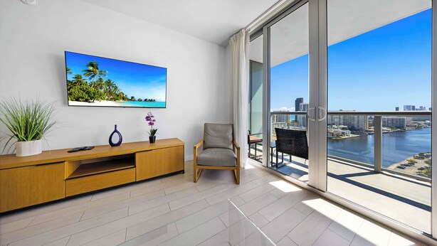 A modern living room with a large window, a wooden entertainment unit, a comfortable chair, and a city view. - Beachwalk Resort 1BR Ocean & Intracoastal View  High Floor 2109 (Hallandale Beach)