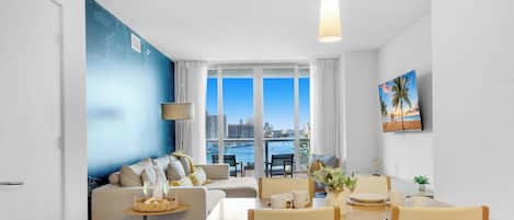 Una sala de estar moderna con sofá, mesa de comedor, sillas y vistas a la ciudad a través de amplias ventanas.