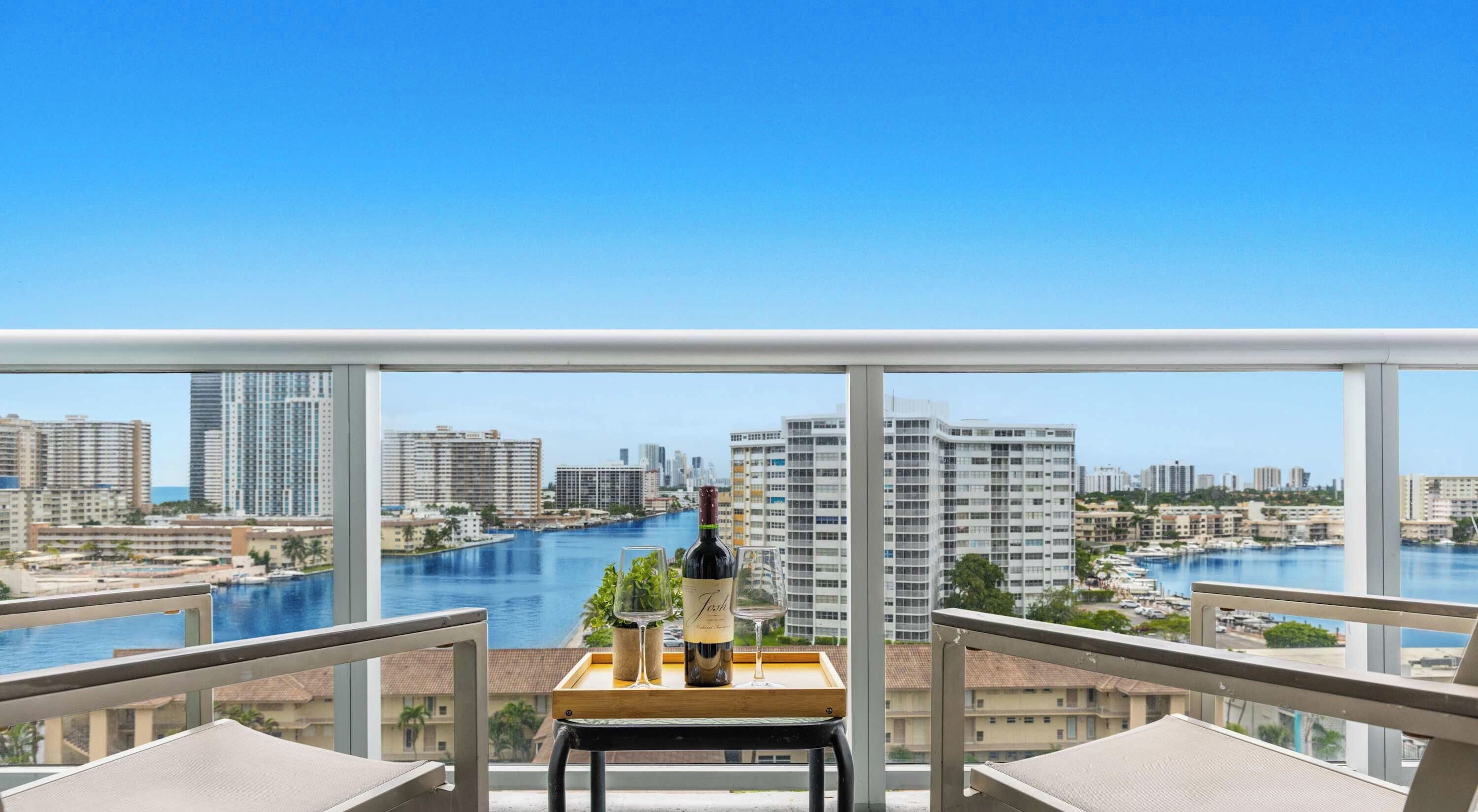 Un balcon avec une table, une bouteille de vin et un verre, donnant sur un paysage urbain avec une rivière et des immeubles de grande hauteur.