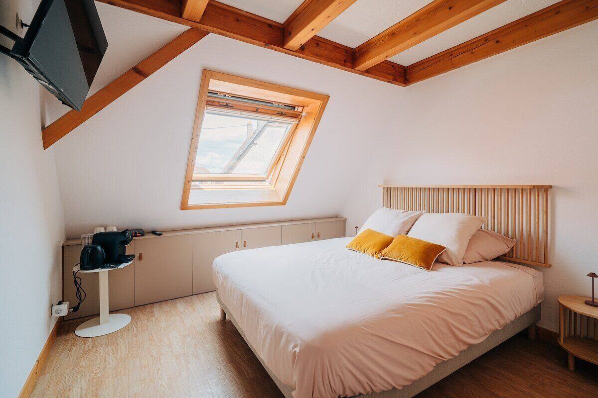Une chambre avec un plafond en pente, une lucarne, un lit avec une literie blanche et deux oreillers, une table de chevet avec une lampe, et un petit tabouret en bois.