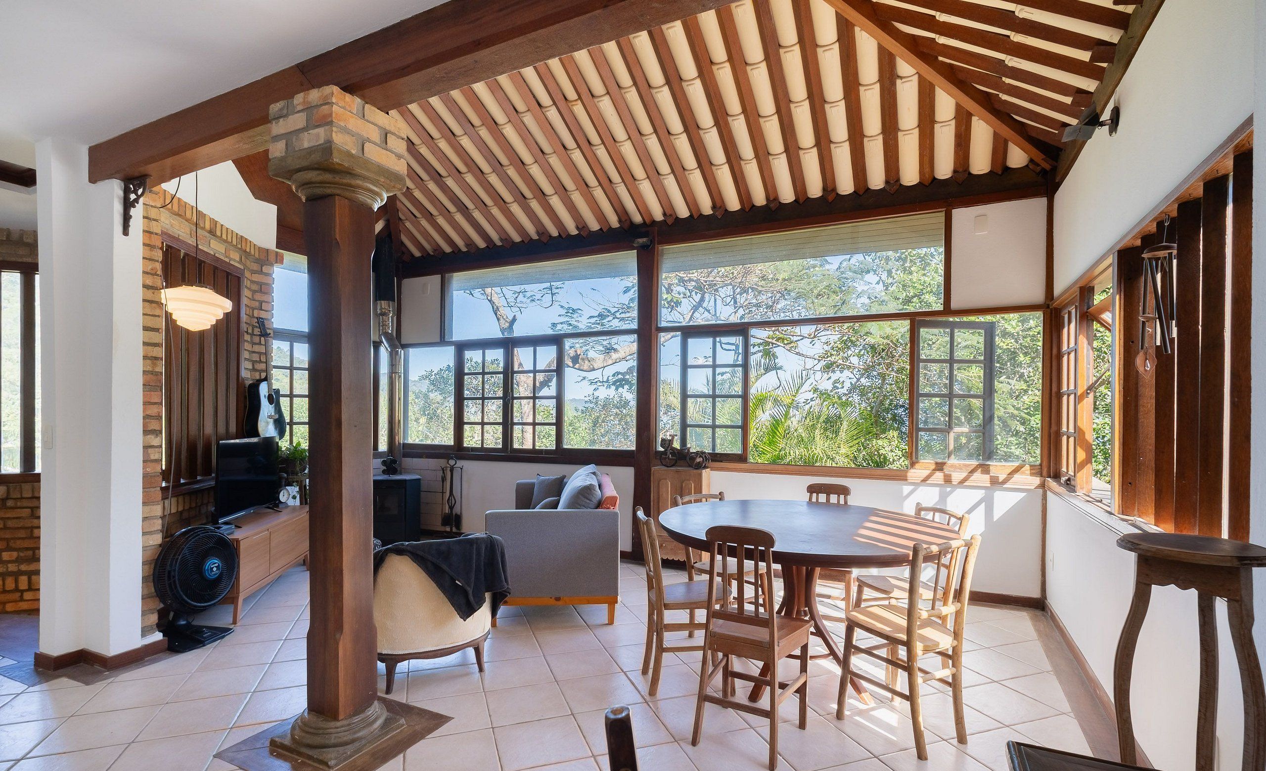 A spacious room with a large dining table, wooden chairs, and a view of greenery through large windows.