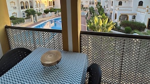 A balcony with a table and chairs, overlooking a pool and buildings with Mediterranean architecture.