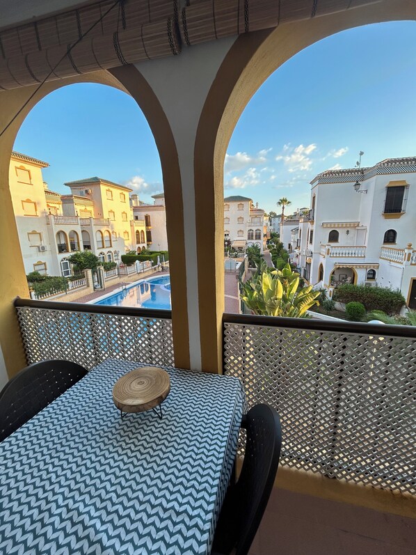 Un balcón con mesa y sillas, con vistas a una piscina y edificios de arquitectura mediterránea.