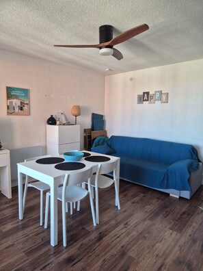 A room with a ceiling fan, a blue sofa, a white table with chairs, and a small cabinet.