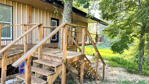 Una cabaña de madera con una escalera de madera que lleva a una terraza, rodeada de árboles y follaje.