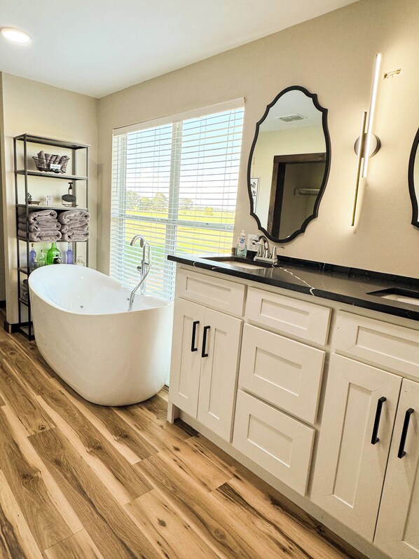 A modern bathroom with a freestanding bathtub, wooden flooring, and a large window providing natural light.