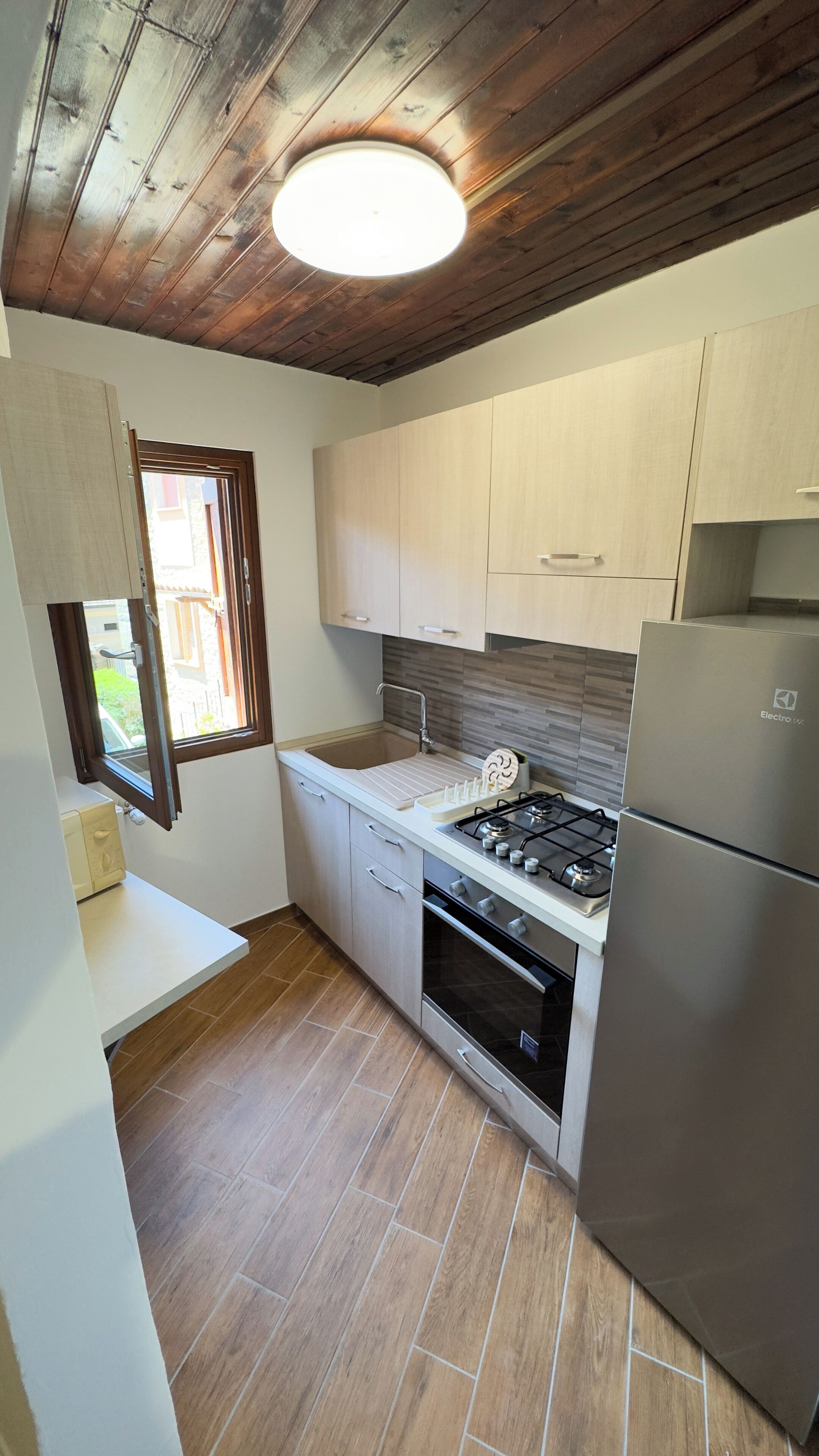A compact kitchen with wooden ceiling beams, a built-in oven, a stainless steel refrigerator, and a window with a view of the outside.