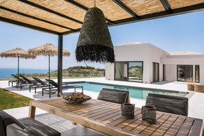 Un área de piscina exterior moderna con sillones reclinables, una mesa de madera y una lámpara colgante.
