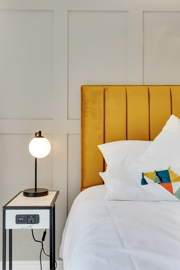 A neatly made bed with a white comforter and a yellow headboard, a bedside table with a lamp, and a power outlet.