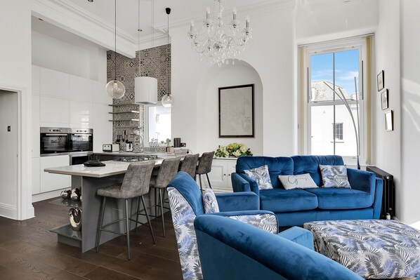 Une cuisine moderne avec des meubles blancs, un îlot central et des tabourets de bar. On y trouve un canapé bleu avec des coussins à motifs et un lustre au-dessus.