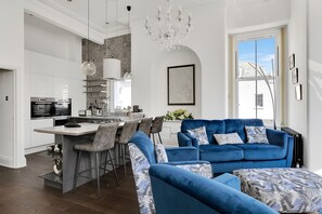 Une cuisine moderne avec des meubles blancs, un îlot central et des tabourets de bar. On y trouve un canapé bleu avec des coussins à motifs et un lustre au-dessus.