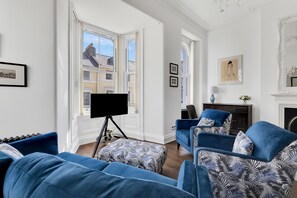 A living room with a large window, a flat-screen TV on a tripod, and blue upholstered furniture.