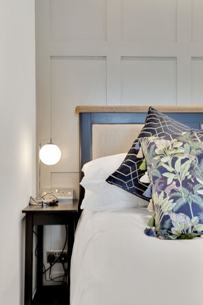 A neatly made bed with white linens and decorative pillows, a bedside lamp, and a power outlet on a nightstand.