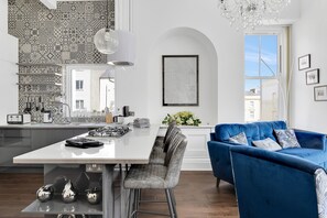 A modern kitchen with a white island, grey bar stools, and a blue sofa with patterned cushions.