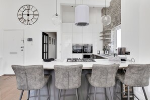 Une cuisine moderne avec des meubles blancs, un îlot central doté de tabourets, une grande horloge et des luminaires suspendus.