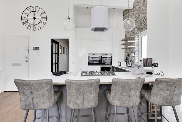 A modern kitchen with white cabinets, a central island with bar stools, a large clock, and pendant lights.
