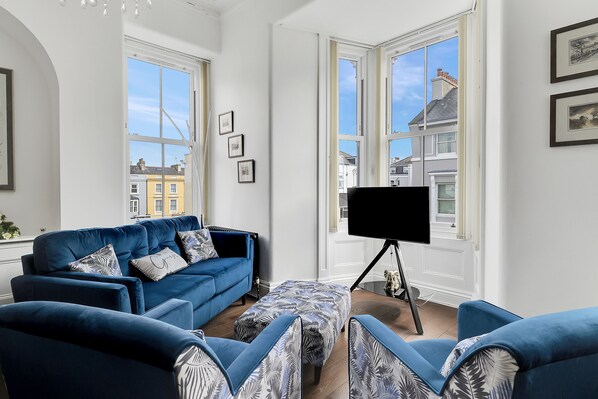 Un salon avec un canapé bleu, un téléviseur à écran plat posé sur un trépied et de grandes fenêtres donnant sur des immeubles.