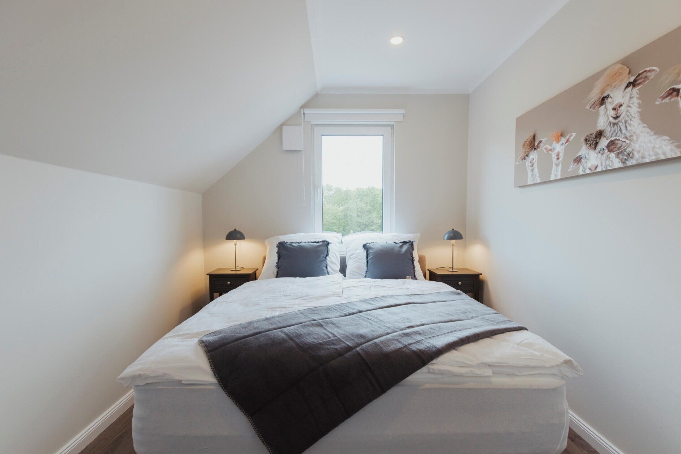 A bedroom with a large bed, two bedside tables, a window, and a wall-mounted painting.