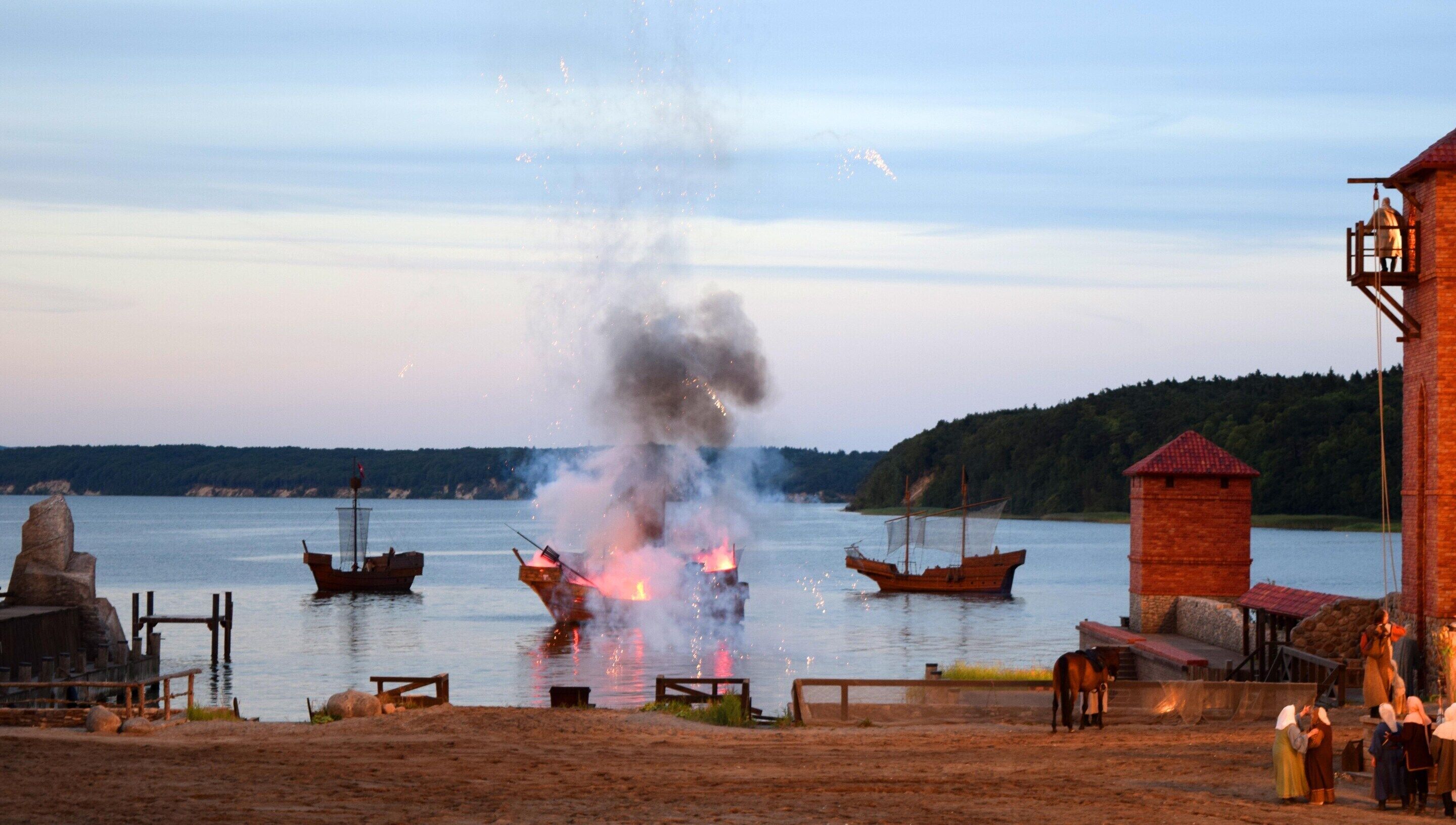 A historical reenactment scene with boats and a wooden structure by the water.