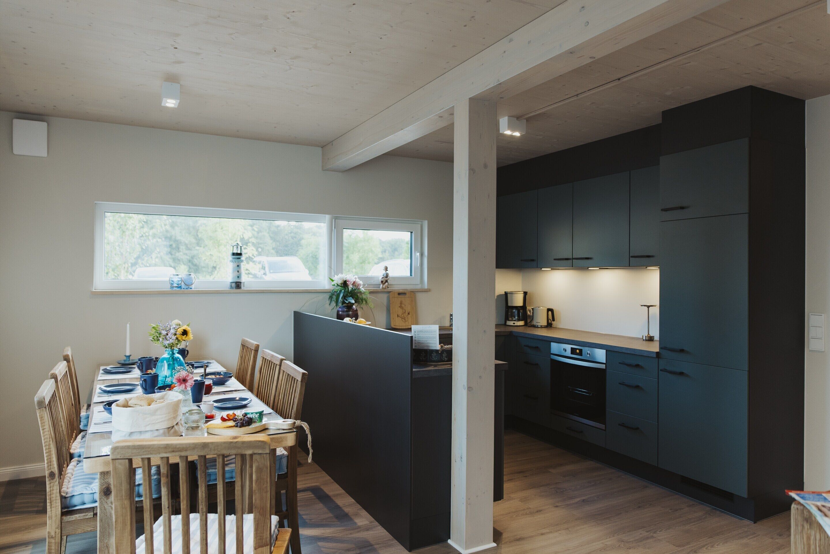 A modern kitchen with dark cabinetry, a dining area with wooden chairs and a table set for four, and a view of the outdoors through large windows.