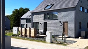 A modern two-story house with a sloped roof, large windows, and a visible outdoor area with a wooden deck and a small garden. - Skandinavisch Eingerichtetes Familienferienhaus (Sagard)